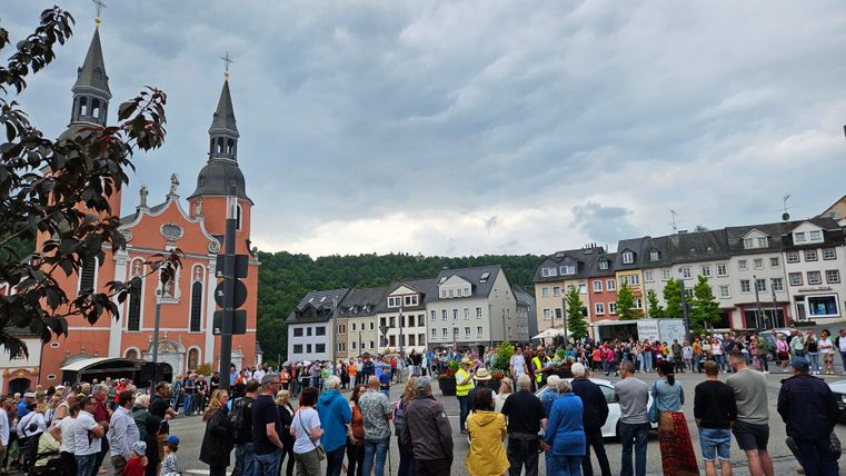 Une place animée avec une église en arrière-plan et beaucoup de gens. Le ciel est nuageux et l'atmosphère semble festive.