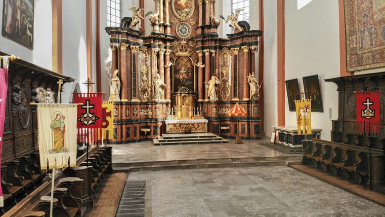 Ein alterswürdiger Kirchenraum mit einem aufwendigen Altar und bunten Fahnen. Die Wände sind hell, und Licht strömt durch die Fenster.