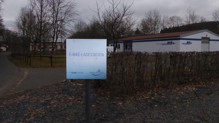 Eine E-Bike-Ladestation ist deutlich sichtbar. Im Hintergrund ist ein Gebäude mit einer grauen Fassade und kahlen Bäumen.