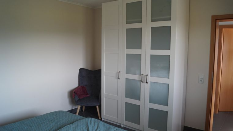 Ein einfaches Schlafzimmer mit einem Bett und einem grauen Sessel. Der weiße Kleiderschrank hat Glastüren und steht an der Wand.