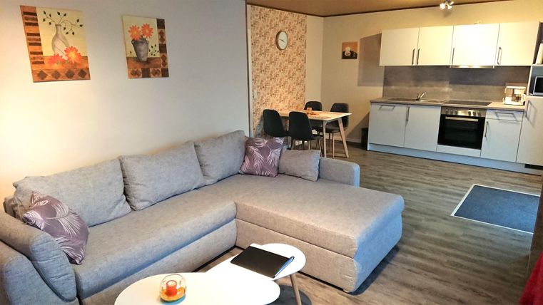 Ein modernes Wohnzimmer mit einer grauen Couch und einem kleinen Tisch. Im Hintergrund ist eine offene Küche mit Essbereich zu sehen.