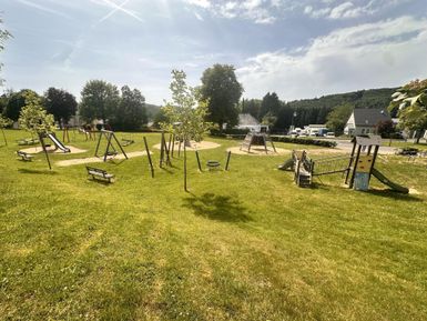 Ein Spielplatz mit verschiedenen Spielgeräten und viel grünem Gras. Im Hintergrund sind Bäume und einige benutzerfreundliche Sitzgelegenheiten zu sehen.