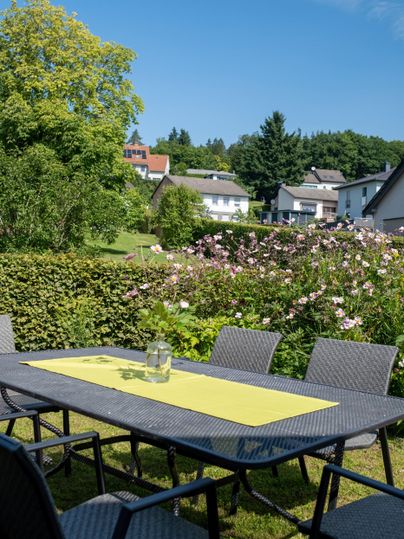 Ein schöner Garten mit einem Tisch und Stühlen. Im Hintergrund sind gepflegte Blumen und moderne Häuser zu sehen.