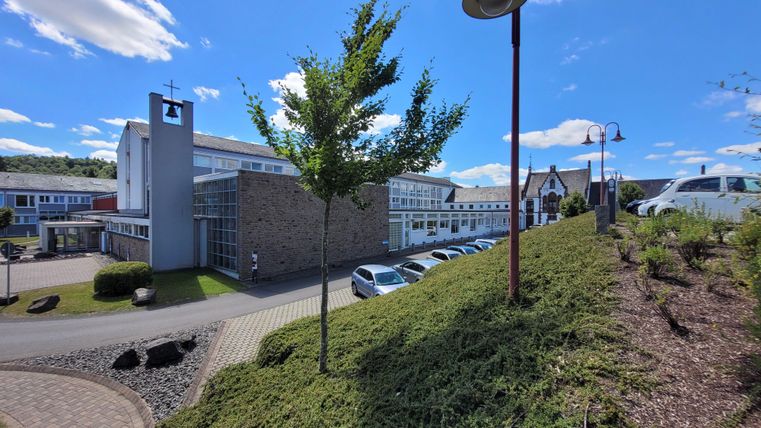 Ein modernes Gebäude mit großen Fenstern und einer grünen Landschaft im Vordergrund. Der Himmel ist klar und blau, was eine freundliche Atmosphäre schafft.