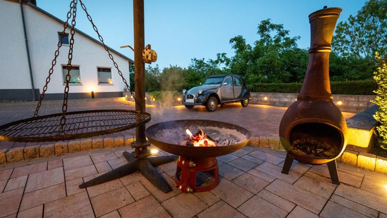 Ein gemütlicher Außenbereich mit einem Feuerplatz, einem Grill und einer Feuerstelle. Im Hintergrund steht ein Auto und die Umgebung ist sanft beleuchtet.