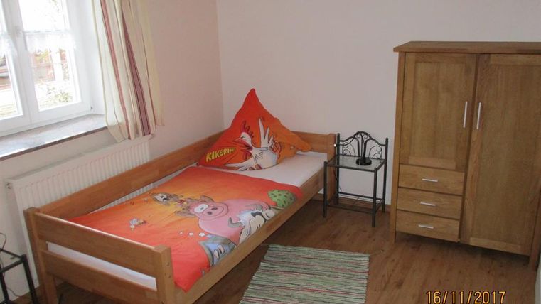 Ein einfaches Schlafzimmer mit einem Einzelbett und bunten Bettwäsche. Neben dem Bett steht ein Holzkasten und ein kleiner Stuhl.