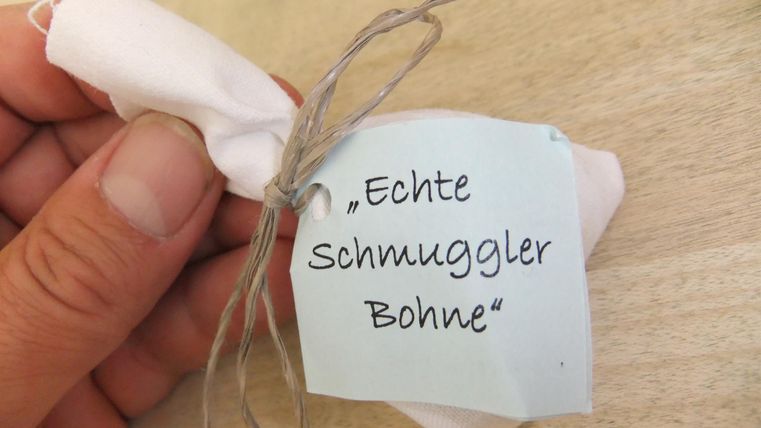 Eine Hand hält ein kleines Päckchen mit einem Aufkleber, der "Echte Schmuggler Bohne" sagt. Das Päckchen ist mit einem Stück Schnur gebunden und sieht aus wie ein Geschenk.