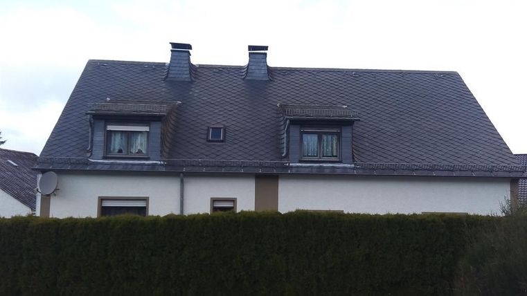 Ein zweigeschossiges Haus mit einem schrägen Dach und zwei kleinen Dachfenstern. Im Vordergrund befindet sich eine grüne Hecke.