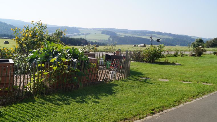 Ein schöner Garten mit Gemüsebeeten und einem Bienenstock. Im Hintergrund erstrecken sich sanfte Hügel unter einem klaren Himmel.
