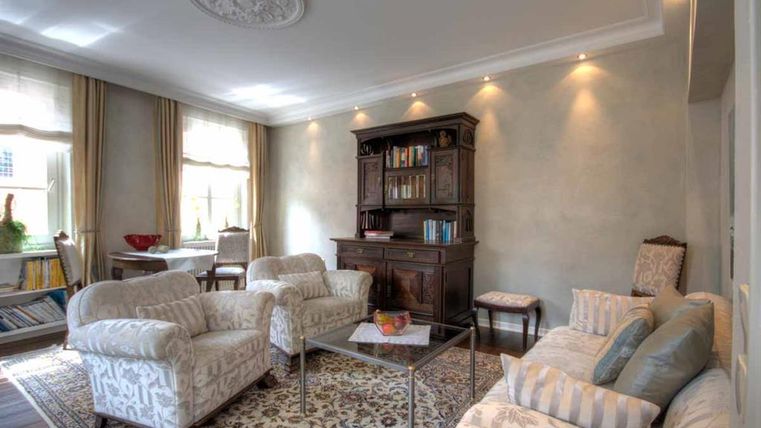 Ein gemütliches Wohnzimmer mit eleganten Möbeln und einem stilvollen Bücherregal. Die großen Fenster sorgen für viel Tageslicht und eine einladende Atmosphäre.