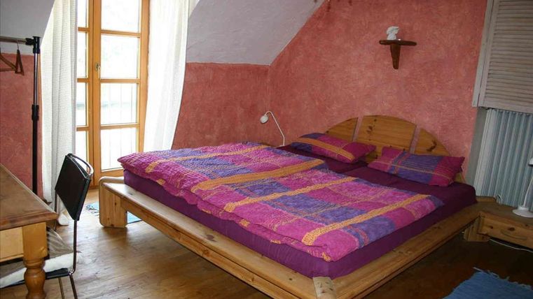 Ein gemütliches Schlafzimmer mit einem großen Holzbett und bunten Bettdecken. Viel Tageslicht strömt durch die Fenster, die einen warmen, einladenden Raum schaffen.