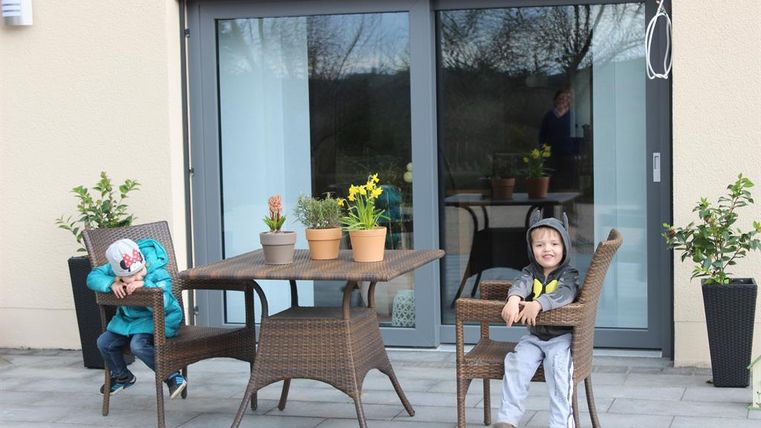 Zwei Kinder sitzen auf Stühlen an einem Tisch auf einer Terrasse. Im Hintergrund sind Pflanzen und große Fenster zu sehen.