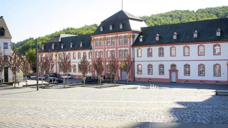 Ein historisches Gebäude mit einer eleganten Fassade steht auf einem gepflasterten Platz. Im Hintergrund sind sanfte Hügel zu sehen.