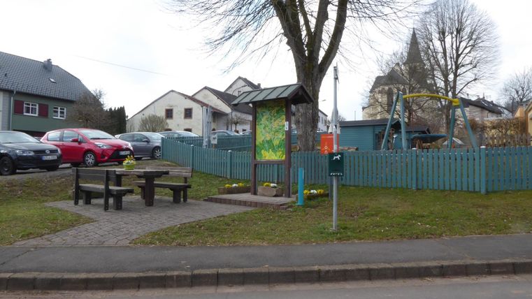 Ein kleiner Spielplatz mit einer Schaukel und einem Tischbereich. Im Hintergrund sind ein paar Autos und Häuser sichtbar.
