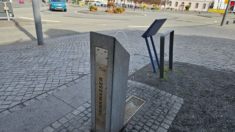 Ein Trinkwasserbrunnen aus Stein steht auf einem gepflasterten Platz. Im Hintergrund sind Autos und ein Schild sichtbar.