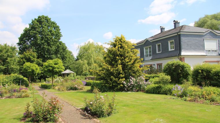 Ein schöner Garten mit bunten Blumen und gut gepflegtem Gras. Im Hintergrund steht ein modernes Haus unter blauem Himmel.