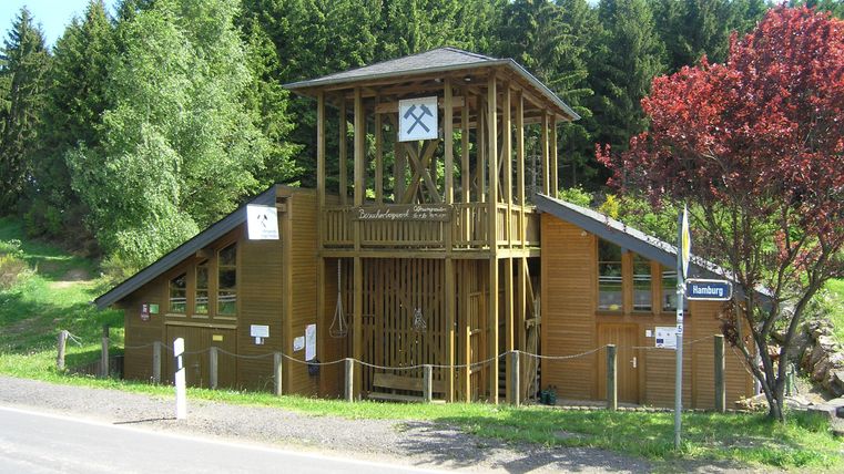 Ein modernes Holzgebäude umgeben von Bäumen. Es hat einen markanten Turm mit einer Uhr und liegt an einer Straße.
