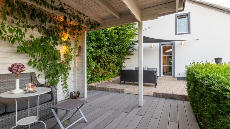 Eine gemütliche Terrasse mit Gartenmöbeln und grünen Pflanzen. Im Hintergrund ist ein modernes Haus mit großen Fenstern zu sehen.