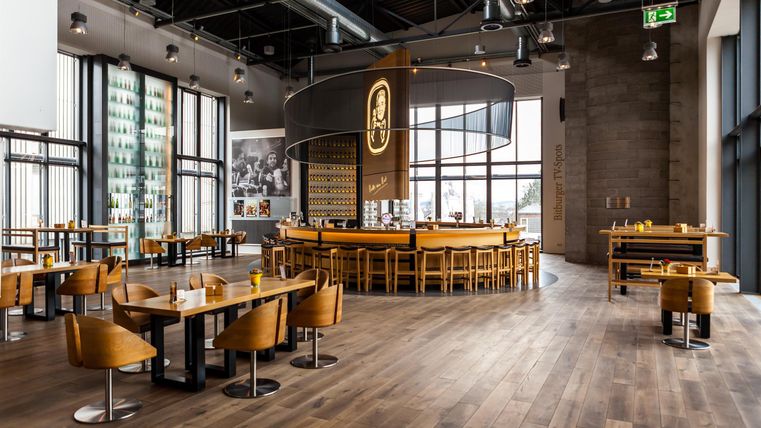 Een modern restaurant met een centrale bar en houten meubels. Grote ramen zorgen voor veel daglicht en een uitnodigende sfeer.
