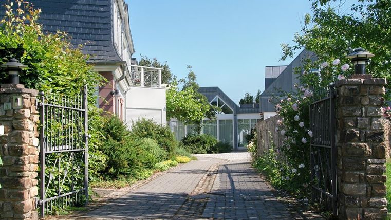 Ein schöner Zugang zu einem gepflegten Garten mit Bäumen und Blumen. Die gepflasterte Auffahrt führt zu einem modernen Haus.