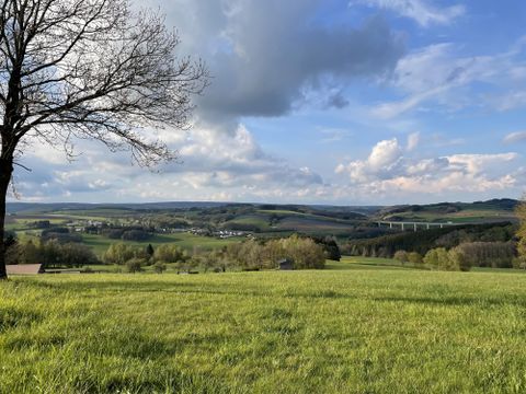 Eine weitläufige grüne Landschaft mit sanften Hügeln und einem blauen Himmel. Im Hintergrund sind Wolken und eine kleine Ortschaft zu sehen.