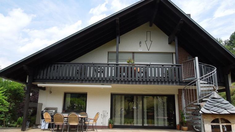Ein modernes Haus mit einem großen Balkon und einer Treppe. Davor befindet sich ein gemütlicher Außenbereich mit Gartenmöbeln.