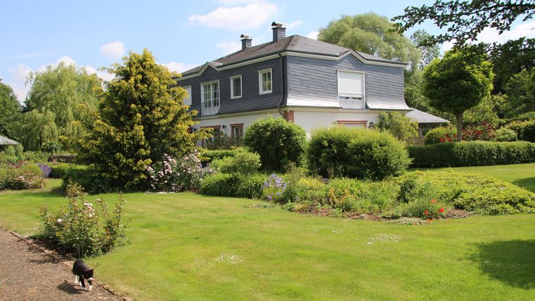 Ein schönes Haus umgeben von grünem Garten und Bäumen. Der Himmel ist klar und sonnig.