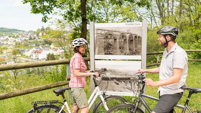 Zwei Radfahrer stehen vor einem Infopunkt am Maare-Mosel-Radweg mit historischen Fotos des Viadukts Daun.