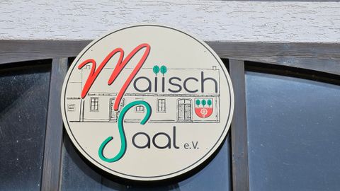Un panneau rond avec l'inscription "Maiisch Saal e.V." en lettres colorées. En arrière-plan, un bâtiment est suggéré.