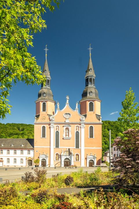 Eine beeindruckende Kirche mit zwei Türmen und einer auffälligen, orangefarbenen Fassade. Umgeben von grünem Laub und einem klaren blauen Himmel.
