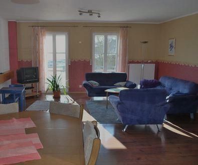 Ein helles Wohnzimmer mit blauen Sofas und einem Tisch. Große Fenster sorgen für viel Tageslicht.