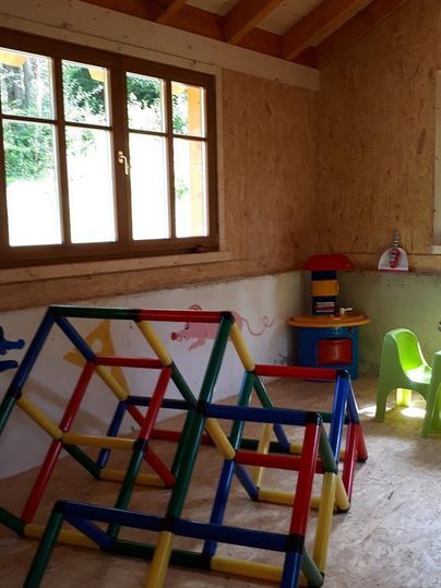 Ein Spielzimmer mit bunten Klettergerüsten und einem Tisch mit Stühlen. Das Zimmer hat große Fenster und eine gemütliche Atmosphäre.