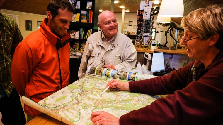 A man in an orange jacket and an older gentleman are listening to a woman who is pointing at a map. The scene takes place in a cozy room decorated with various objects.