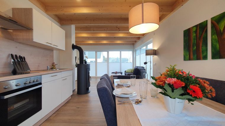 Moderne Küche mit Holzdecke und offenem Wohnbereich. Ein Esstisch mit Blumenarrangement und gemütlicher Sitzecke lädt zum Verweilen ein.