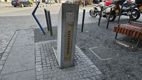 Eine Trinkwasserstelle aus Beton steht auf einem Platz. Im Hintergrund sind Motorräder und Sitzbänke sichtbar.