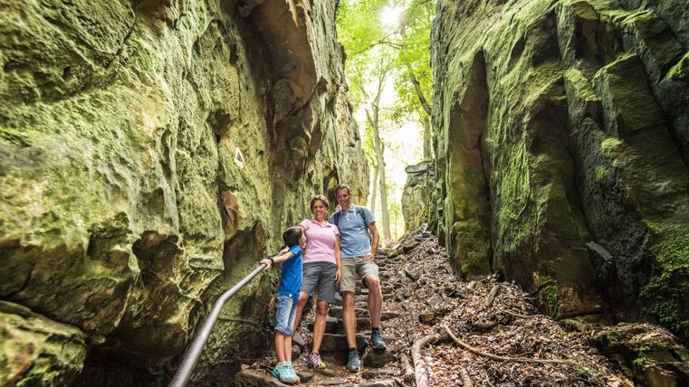 Eine Familie steht auf einem schmalen Weg zwischen hohen Felsen. Die Umgebung ist grün und sonnig, ideal für einen Ausflug in die Natur.