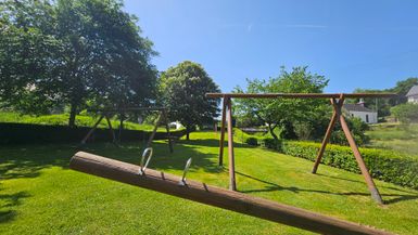 Ein schöner Spielplatz mit Schaukeln in einer grünen Wiese. Der Himmel ist klar und blau, umgeben von Bäumen.