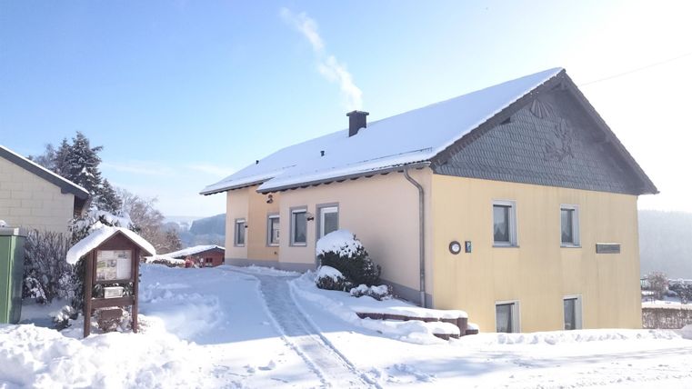Ein Haus im Schnee mit einem rauchenden Schornstein. Die Umgebung ist winterlich und ruhig.