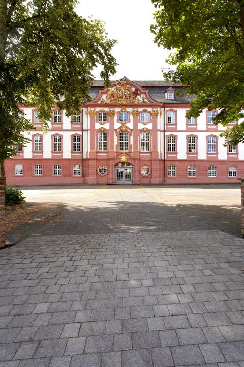 Ein Gebäude mit roter und weißer Fassade, umgeben von Bäumen. Der Weg führt direkt zur Eingangsfront des Hauses.