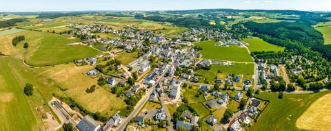 Ein weitläufiges Landschaftsbild mit einem kleinen Dorf, umgeben von grünen Feldern und Hügeln. Die Sonne scheint und der Himmel ist klar.