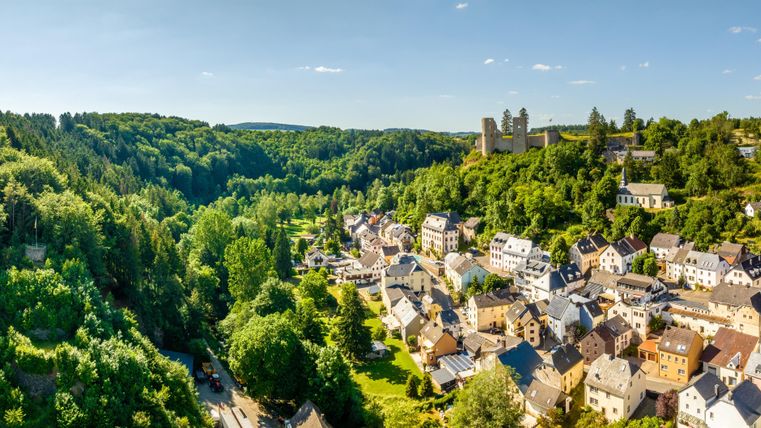 A picturesque landscape with a small village surrounded by lush greenery. In the background, a castle ruin is visible on a hillside.