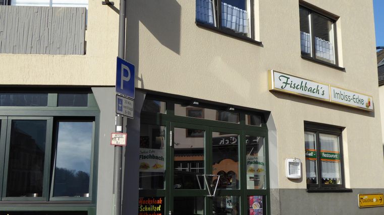 Ein Imbiss mit dem Namen "Fischbach's" in einem hellen Gebäude. Die Fenster sind groß und es gibt eine Markise sowie ein Parkschild vor der Tür.