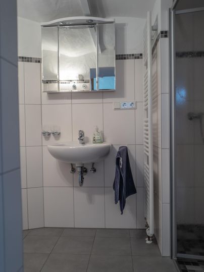 Ein modernes Badezimmer mit einem Waschbecken, einem Spiegel und einer Dusche. Die Wände sind hell gefliest und ein Handtuch hängt am Waschbecken.