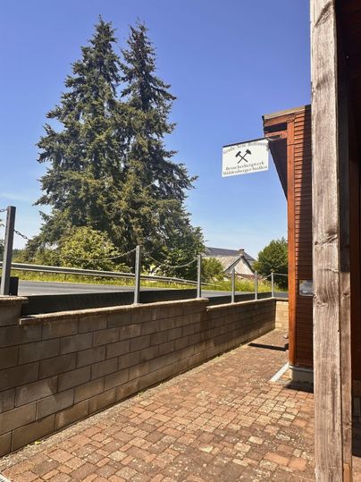Eine schmale Straße mit einem gepflasterten Gehweg und einer Mauer aus Steinen. Im Hintergrund sind hohe Bäume und ein Schild sichtbar.