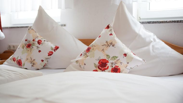 Ein gemütliches Bett mit weißen Kissen und farbenfrohen, blumigen Zierkissen. Das Zimmer hat viel Tageslicht durch die Fenster.