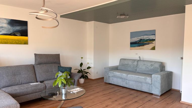 Ein modernes Wohnzimmer mit einer grauen Couch und einem Glastisch. An den Wänden hängen verschiedene Bilder und eine Pflanze steht in der Ecke.