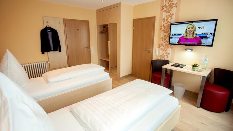 Ein modernes Zimmer mit zwei Einzelbetten und einem Fernseher an der Wand. Es gibt einen Tisch, Stühle und einen Kleiderschrank für persönlichen Stauraum.