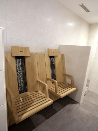 Zwei moderne Holzliegen stehen in einem hellen Raum. Die Wände sind schlicht und hell gefliest.