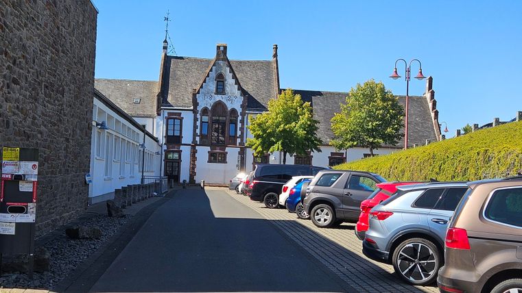 Une étroite rue avec des voitures garées mène à un bâtiment historique. Le temps est ensoleillé et le ciel est bleu.