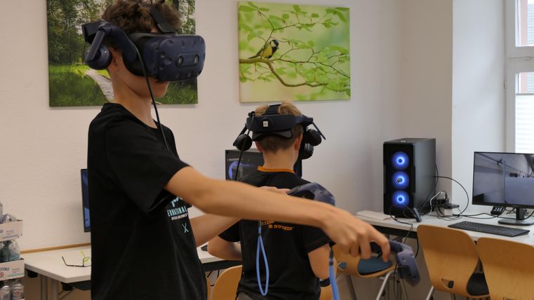 Two children are wearing virtual reality headsets and interacting with the technology. In the background, computers and pictures on the wall are visible.